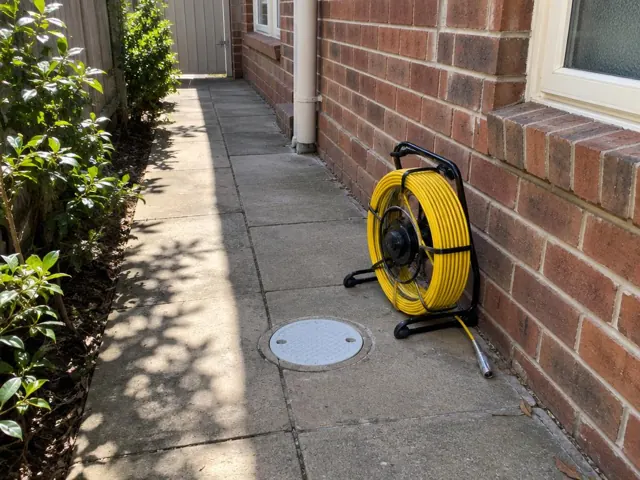 Capped inspection opening beside a coiled drain camera reel on a brick house side path in Artarmon