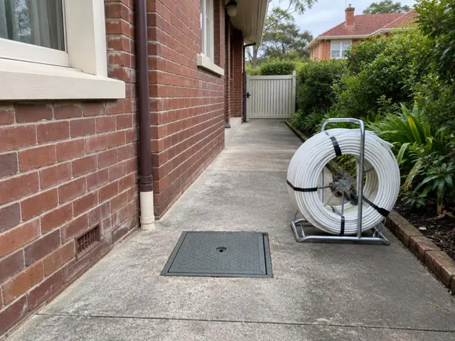 Concrete side path beside a brick house with a flush inspection cover and pipe reel in Lane Cove