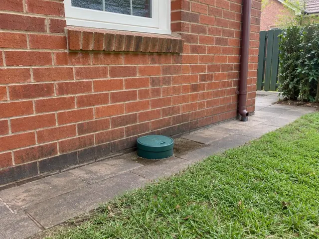 Green capped sewer opening beside a brick house wall and paved path in Artarmon
